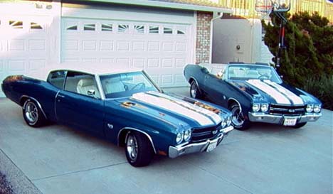 Two blue muscle cars parked in front of a garage.